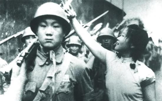 1949年7月6日，上海举行军民大游行，欢天喜地庆解放。一位上海姑娘把鲜花挂到了解放军战士的枪上。