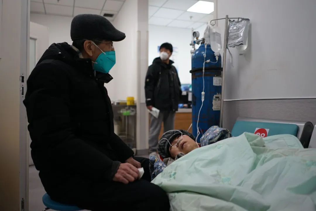 2022年12月27日，北京丰台，一位感染新冠一周后基础病加重的老人临时躺在首都医科大学附属北京佑安医院急诊科的座椅上，一边输液、吸氧，一边等待病床，她的丈夫与儿子陪在身旁。中青报·中青网记者李强/摄